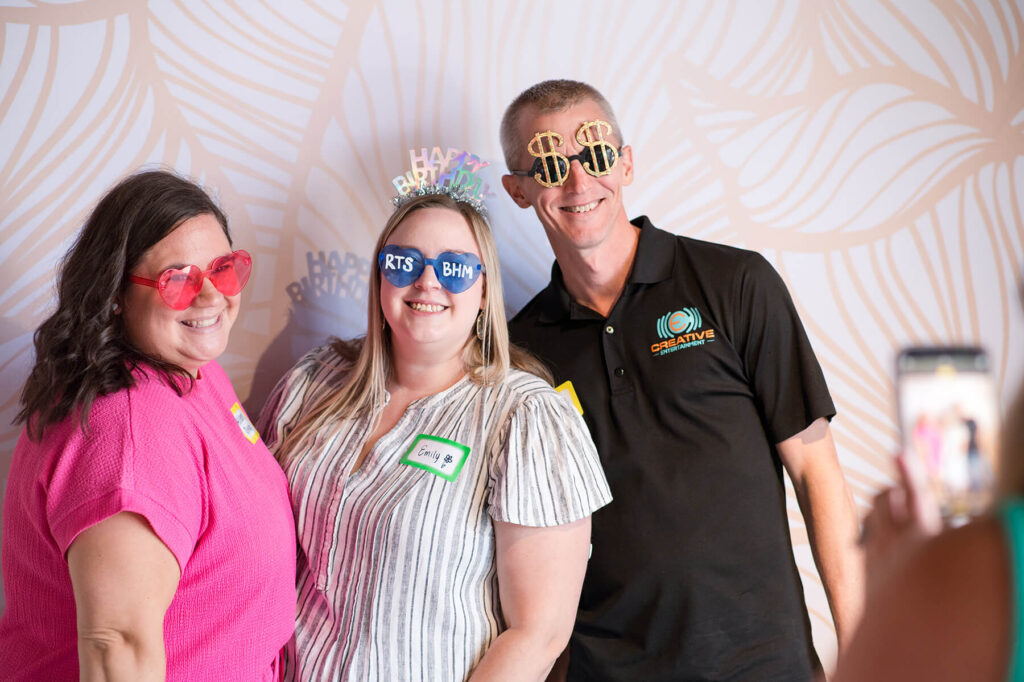 Three Birmingham Rising Tide members wearing celebratory glasses  and happy birthday headbands in front of a photo wall