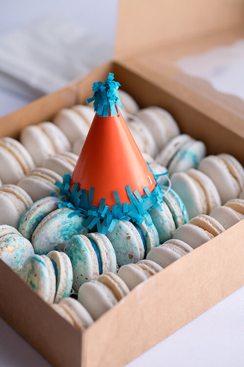 Mini orange and blue party hat on top of a box of macrons