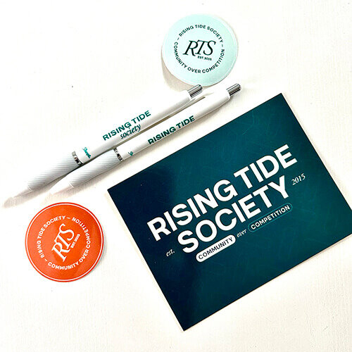 Two pens, two stickers and a postcard that have the Rising Tide Society logo